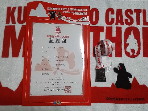 赤だらけで分かりにくいかもしれませんが，フィニッシャータオルと記録証，完走メダルです．記録証を入れるケースももらえます．メダルの形は，熊本城の石垣なので武者返しも分かりますかね？　Tシャツは，まだ手にしていない人がおられるので，あえてアップしませんでしたが，背中に大きく「挑（いどめ）」の筆文字が入っています．でもつい「桃」に見えてしまう気がするのは私だけ･･･